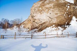 A rock against the blue sky, a man's shadow on white snow in winter, a wooden fence made of logs, a fence for animals on a ranch, a hike in the mountains in winter. photo