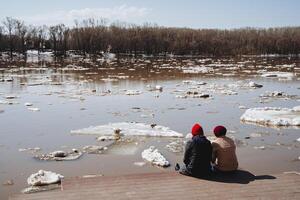 dos personas sentar lado por lado en el río banco, comunicación Entre dos gente, inundación en el río, sucio agua flotante hielo primavera, soleado día en un caminar en el parque, ciudad terraplén. foto