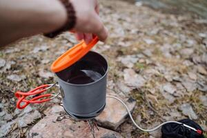 abierto el tapa de el caldera con tu mano, cheque el hirviendo agua en el maceta, el maceta con el tapa, el turista platos, hervir el agua para té en el gas quemador en un cámping en naturaleza. foto