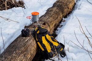 Halt parking in the forest, yellow backpack lies on the snow, burner stands on the log to prepare lunch on a hike, camping equipment, survival in the wild cold photo