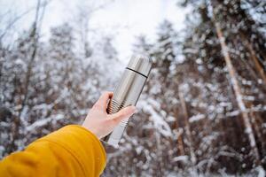 Thermos in hand against the background of the forest. The concept of vacuum objects. Hot tea on a hike in the mountains. Extend your arm up. photo