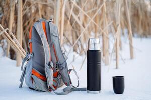 A black thermos of tea stands next to the backpack. Vessel for hot drinks. A small backpack for a hike. Rest on the lake. photo
