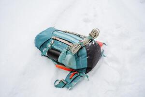A bright hiking backpack lies on the snow. A climbing rope is tied to a backpack. In the side pocket of the thermos. Hiking in winter. Rocks and forests photo