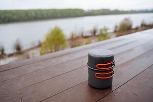 A pot with a lid stands on a table against the background of the river in nature, tourist dishes for eating, compact equipment for hiking, a pot with handles. photo