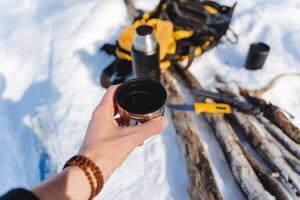 A mug in his hand against the background of hiking equipment. Hot tea in a metal cup. Breakfast in the woods, Survival in nature in winter. Bushcraft. photo