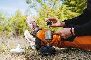 comiendo en el bosque en el abierto aire, Cocinando en un caminata en un gas quemador, manos participación un taza, un tarro de té, un caliente beber, espolvorear sal en alimento, comiendo sublima. foto