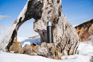 The thermos stands against the background of an old broken tree, the concept of traveling in winter, spring weather, warm sun, a hike in the forest. photo