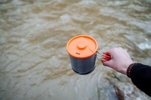 A person holds in his hand a pot with a lid against the background of water, tourist dishes, a pot for eating on a hike. photo