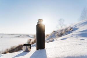 Sunlight glints with rays against the blue sky. A thermos of hot tea stands in the snow. Winter landscape, hiking in the forest, camping equipment. photo