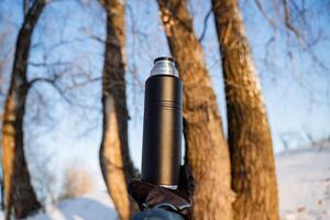 The thermos is black against the background of the forest. Steel bottle for tea. Maximum heat retention of the liquid. Hot tea on a hike. Tableware for traveling in winter. photo