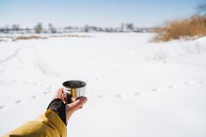 Thermal mug in hand, metal cup for hot tea. Camping on a clear sunny day, Breakfast against the backdrop of snow. photo