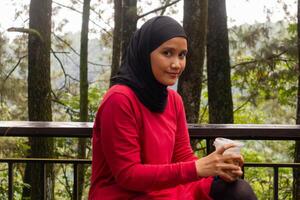 An Asian Muslim woman sitting on the bench and holding a plastic cup of ice coffee with nature or trees background photo