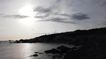 The silhouette of two people walking on the rocks overlooking a small bay in Sardinia at sunset on a cloudy day. video