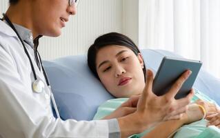 Patient lying in hospital bed looking at digital tablet with doctor talking her health photo