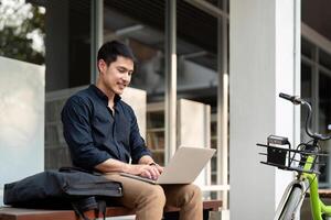 Asian businessman with bicycle using laptop and sitting outside the office building. man commuting on bike go to work. Eco friendly vehicle, sustainable lifestyle concept photo