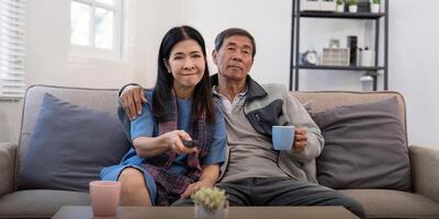 mayor Pareja acecho televisión y sofá en relajarse para película o serie en vivo habitación a hogar. mayor hombre y mujer con café y remoto juntos para cambiando canal y en línea entretenimiento foto