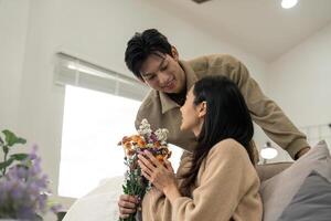 Romantic young asian couple embracing with holding flowers and smiling in living room at home. fall in love. Valentine concept photo