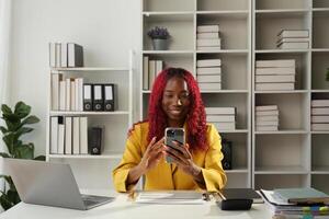 African businesswoman using smartphone phone app for playing games, shopping online, and ordering delivery while working on a laptop computer in the office photo