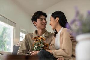 Romantic young asian couple embracing with holding flowers and smiling in living room at home. fall in love. Valentine concept photo