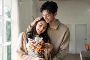 romántico joven asiático Pareja abrazando con participación flores y sonriente en vivo habitación a hogar. otoño en amor. enamorado concepto foto