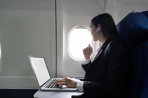 exitoso joven asiático negocio mujer se sienta en avión cabina avión y trabajando desde en cualquier lugar. volador a primero clase foto