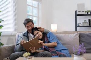 asiático mayor Pareja leyendo libro diario juntos, sentado en acogedor sofá en hogar interior. contento Jubilación actividad estilo de vida a hogar foto