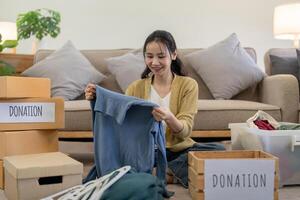 mujer asiático participación donación caja lleno con ropa y Seleccione ropa. concepto de donación y ropa reciclaje. Ayudar pobre personas foto