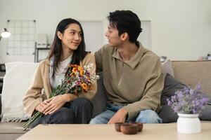 Romantic young asian couple embracing with holding flowers and smiling in living room at home. fall in love. Valentine concept photo