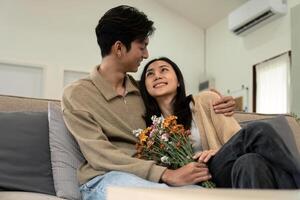 Romantic young asian couple embracing with holding flowers and smiling in living room at home. fall in love. Valentine concept photo