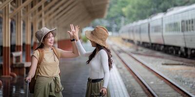 dos mujer son contento mientras de viaje a el tren estación. excursión concepto. dos asiático mujer mejor amigo viaje alto cinco en tren calendario en ferrocarril estación foto