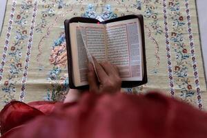 un musulmán mujer sentado en un oración estera y leyendo el Corán con indonesio Traducción foto
