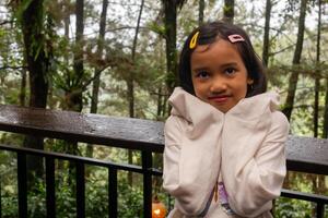 An Asian little girl in long sleeve shirt standing and posing on the balcony with nature or trees background at rainy days photo