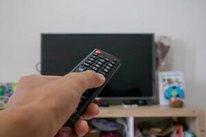 Male hand holding TV remote control directed on the television inside the room photo