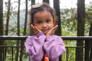 An Asian little girl with curly hair in purple long sleeve shirt posing adorable with trees or nature background photo