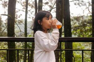 un asiático pequeño niña en pie y Bebiendo un el plastico taza de hielo té en el balcón con naturaleza o arboles antecedentes foto