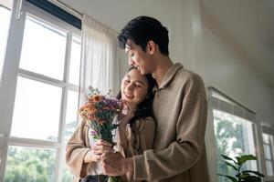 Romantic young asian couple embracing with holding flowers and smiling in living room at home. fall in love. Valentine concept photo