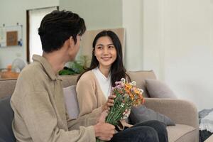 Romantic young asian couple embracing with holding flowers and smiling in living room at home. fall in love. Valentine concept photo