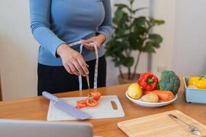 joven mujer asiático medida su cintura en el cocina el con vegetales y frutas concepto de sano comiendo y dieta foto