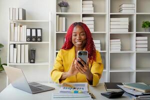 African businesswoman using smartphone phone app for playing games, shopping online, and ordering delivery while working on a laptop computer in the office photo