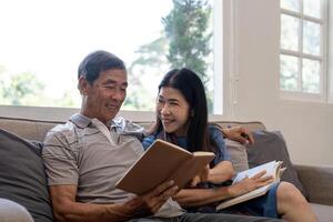 asiático mayor Pareja leyendo libro diario juntos, sentado en acogedor sofá en hogar interior. contento Jubilación actividad estilo de vida a hogar foto