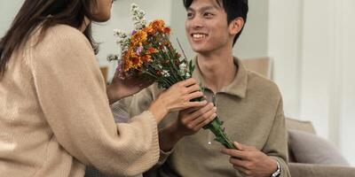 Romantic young asian couple embracing with holding flowers and smiling in living room at home. fall in love. Valentine concept photo