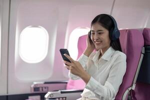 Cheerful Asian woman passenger in headphone watching online movie during intercontinental flight in cabin of aircraft, happy traveler using wifi connection on board photo