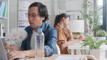 Two Asian workers are unhappy working in hot temperatures at desk in small business office in summer, uncomfortable with no air conditioner, blowing wind with fans, drinking water, and sweating. video