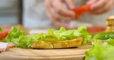 Female chef in the kitchen of the house collects a sandwich. Making a sandwich with ham, salad, ripe tomato slices and pieces of cheese. Fast food concept. Ultra 4K video