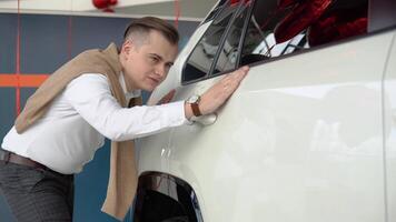 Guy touches the side door of a shiny white car. Young man looking carefully at car in car dealership video