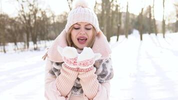 Young woman in a soft pink down jacket, white hat, mittens and scarf blowing on the snow on a background of snowy landscape video