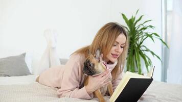 un' giovane donna nel un' accogliente polvere rosa maglione e bianca calze autoreggenti dire bugie su il letto con sua cane e legge un' libro video
