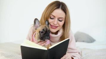 A young woman in a cozy powder pink sweater and white stockings lying on the bed with her dog and a book in her hands looks to the camera video