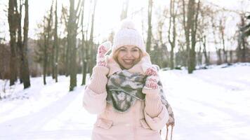 Young woman in a soft pink down jacket, white hat, mittens and scarf smiling at the camera video