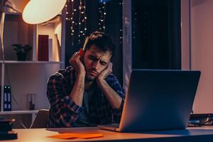 AI generated Young tired male programmer sitting in the office in front of a laptop, lamp light, diffused background photo